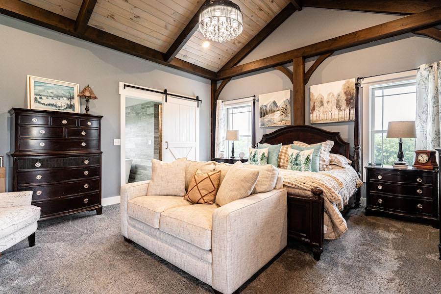 Photo of master bedroom with wood ceiling and dark wood support beams. a sliding barn door covers a glass door leading to an outdoor porch area