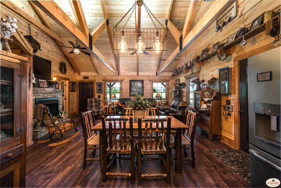 Photo of dining space and living room of log home with exposed wood ceiling and wide a-beams, log exposed walls and stone fireplace