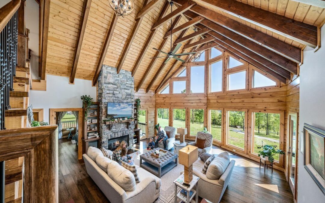 Photo of great room of log home taken from stairs leading to the loft area of the home. The great room has geometric windows angling outward. The ceilings are exposed wood with darker wood support beams. A two story stone fireplace on an interior wall provides a privacy area of living space in the great room