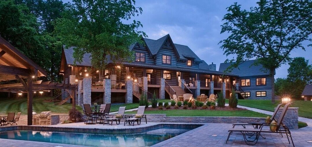 Photo of large log home estate home taken from the l-shaped pool toward the rear of the home with an extensive porch. The pool has a covered a-frame wood structure and two outbuildings can be seen on the property including a two story guest cottage and work shop