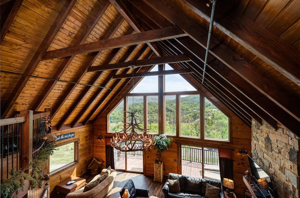 Photo of great room of log home taken from loft showing geometric windows across the exterior wall with glass doors leading to a deck area. The ceiling is dark wood a-frame supporting wood ceiling